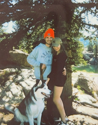 Two people and a dog standing in a forested area with trees and rocks in the background.