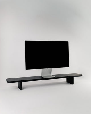 Monolith Desk Shelf in black ash — solid hardwood monitor riser with angled legs and rounded edges, styled with an Apple Studio Display