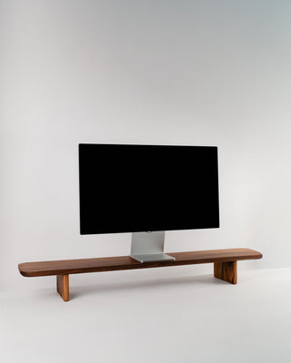Monolith Desk Shelf in walnut — solid hardwood monitor riser with angled legs and rounded edges, styled with an Apple Studio Display