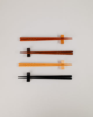 Overhead flat lay of four pairs of chopsticks resting on solid hardwood chopstick rests in cherry, Indian laurel, maple, and black, stacked vertically on a light gray background.