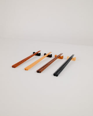 Four pairs of chopsticks resting on solid hardwood chopstick rests, showing all four wood options — Indian laurel, walnut, cherry, and maple — fanned out on a light gray background.