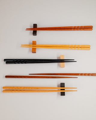 Overhead flat lay of five pairs of chopsticks resting on solid hardwood chopstick rests in walnut, cherry, maple, and Indian laurel, arranged diagonally across a light gray background.