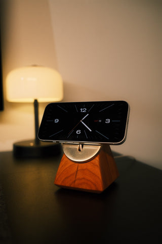 Smartphone on a wooden stand with digital clock display on a table, illuminated by a lamp in the background.